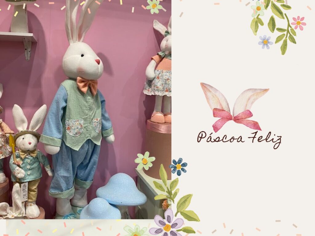 Dois coelhos de pelúcia decorativos, um deles vestindo calça azul, ao lado de um cogumelo azul e branco. A imagem possui um fundo rosa com ilustração de orelhas de coelho e a frase "Páscoa Feliz".