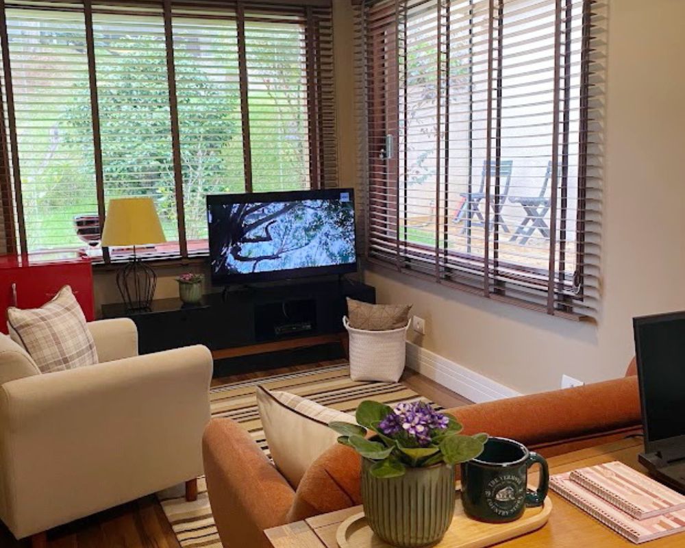 Sala de estar e escritório integrados com janelas amplas, persianas de madeira e vista para o jardim externo.